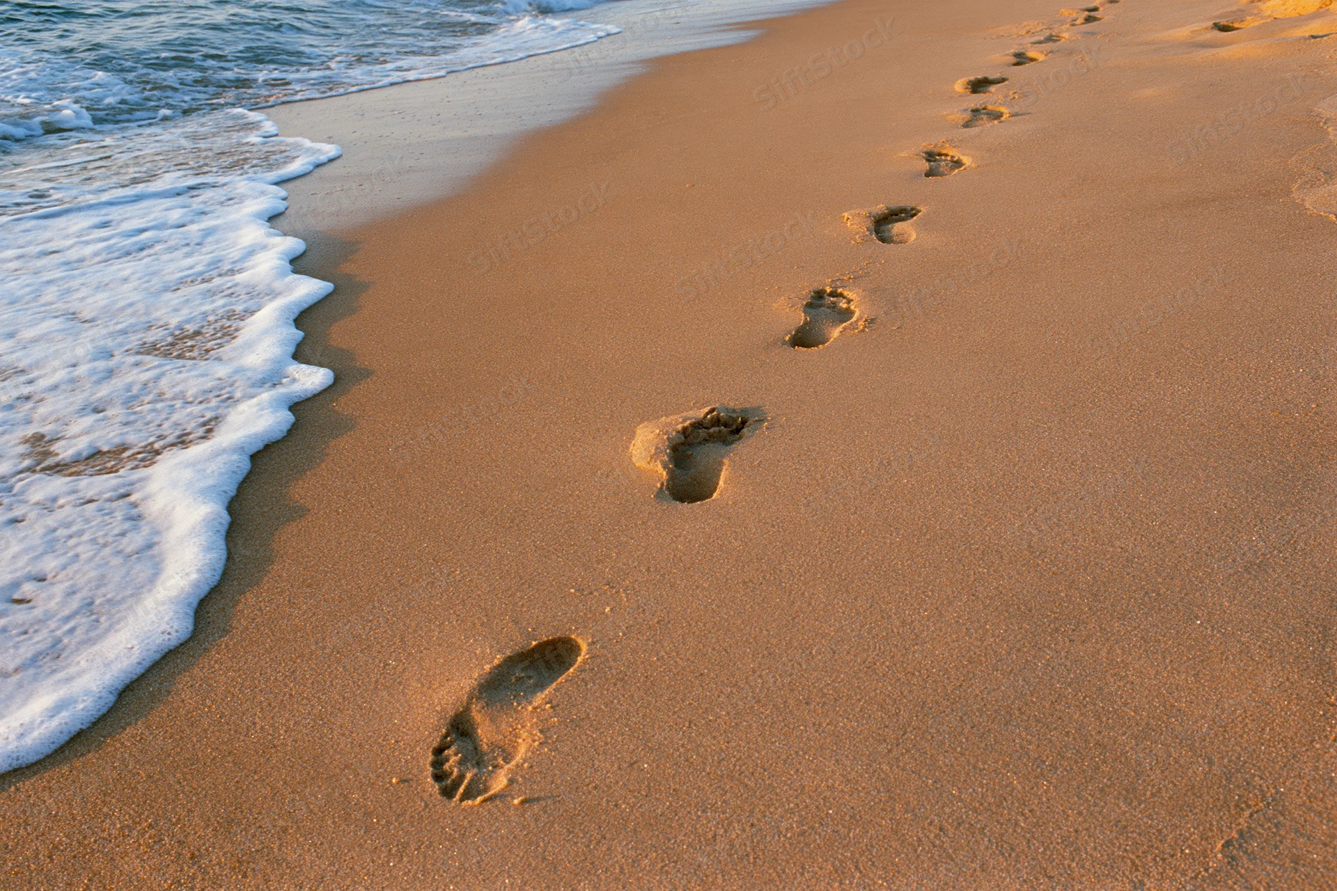 Footprints in the sand along the shoreline with gentle waves at sunset