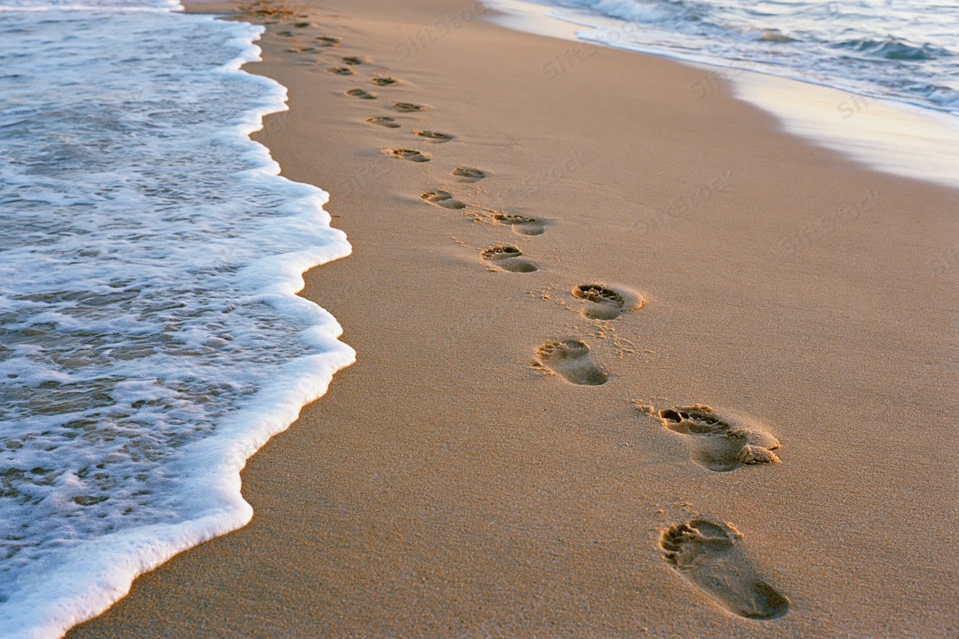 Footprints in the sand along the shoreline with gentle waves approaching at sunrise