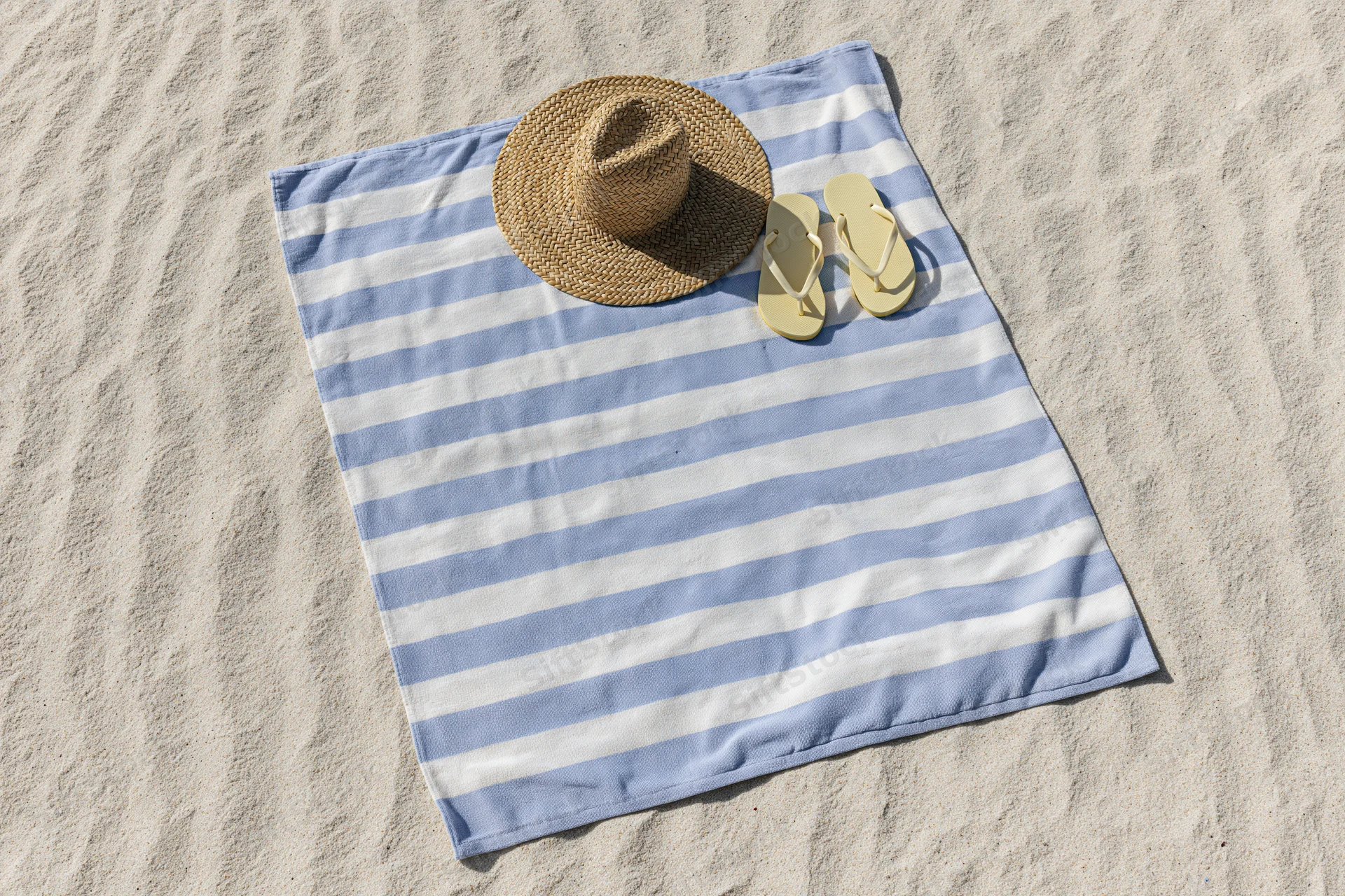 Blue and white striped towel with straw hat and flip flops laid on sandy beach