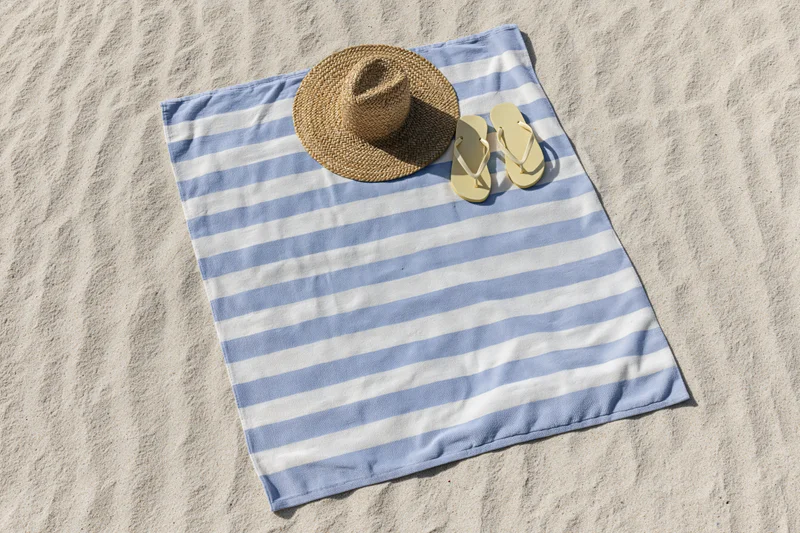 Blue and white striped towel with straw hat and flip flops laid on sandy beach