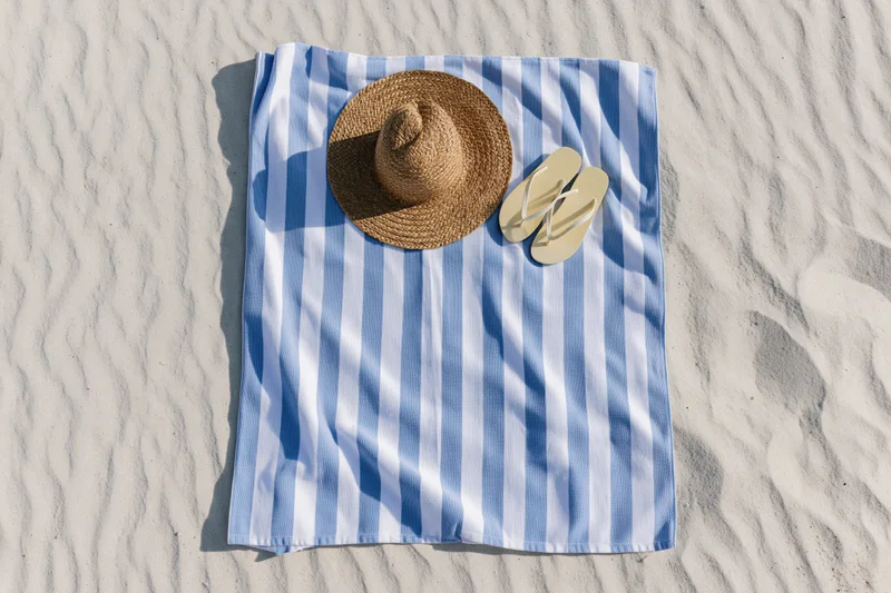 Blue and white striped towel with straw hat and flip flops laid on sandy beach