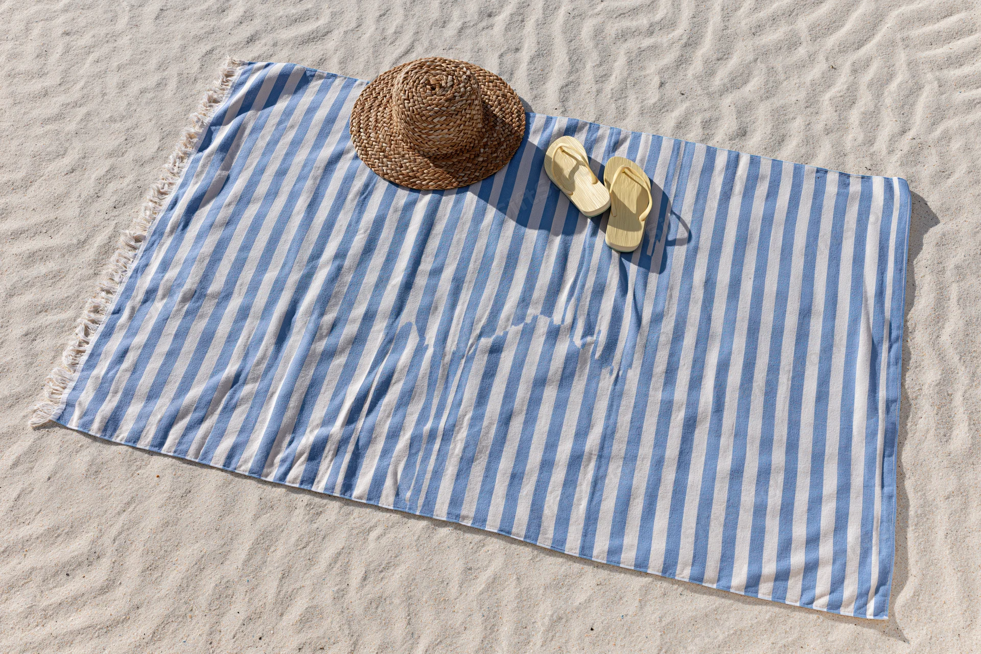 Blue and white striped towel with straw hat and flip flops on sandy beach