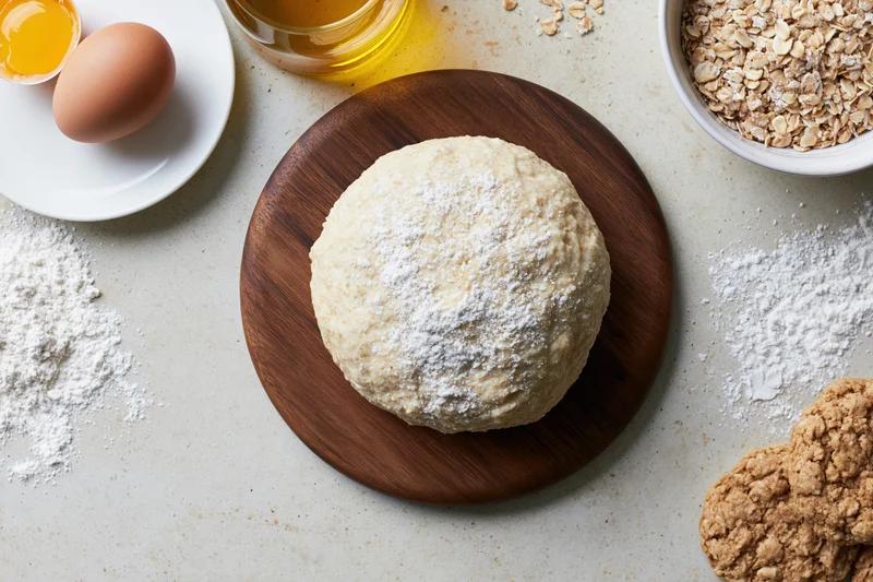 Round ball of dough dusted with flour on wooden board surrounded by baking ingredients on countertop