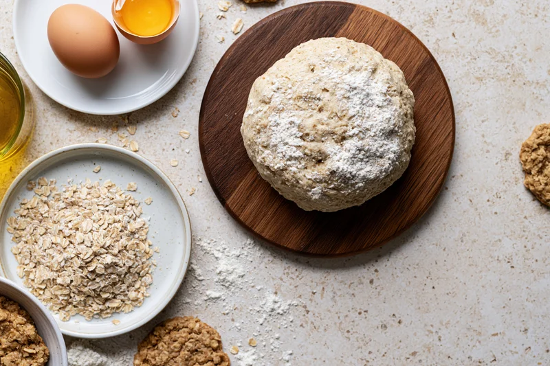 Round dough ball dusted with flour on wooden board surrounded by oats, eggs and baking ingredients on countertop