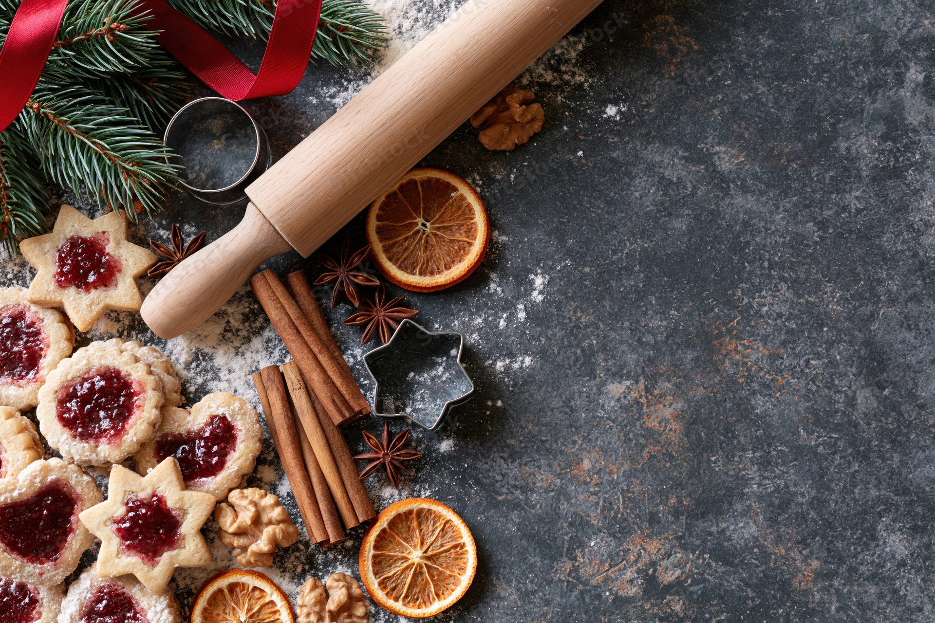 Star-shaped cookies with jam and spices arranged on dark countertop with rolling pin and pine branches for christmas baking