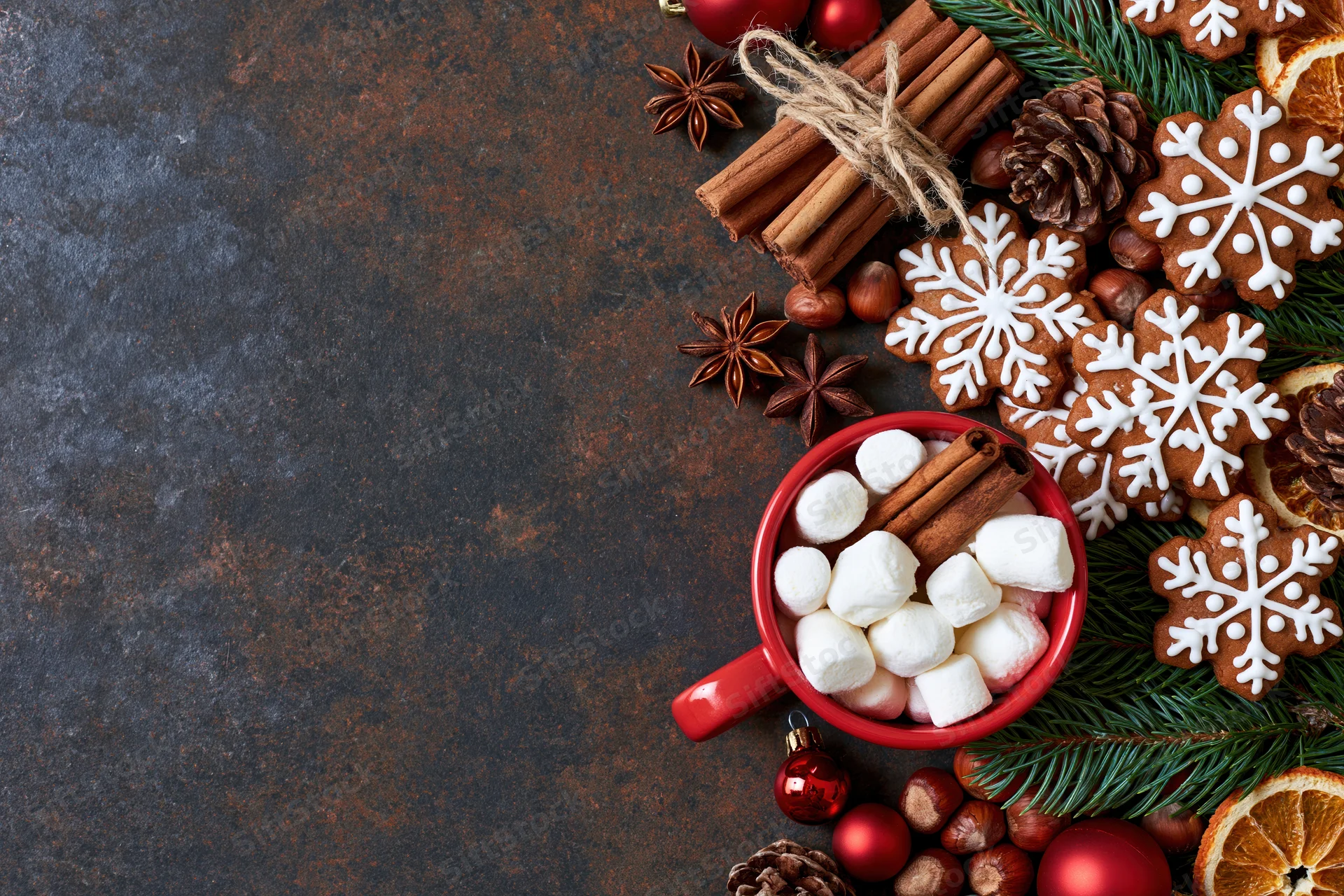 Gingerbread cookies and hot chocolate with marshmallows arranged on a dark textured surface with pine branches and holiday ornaments