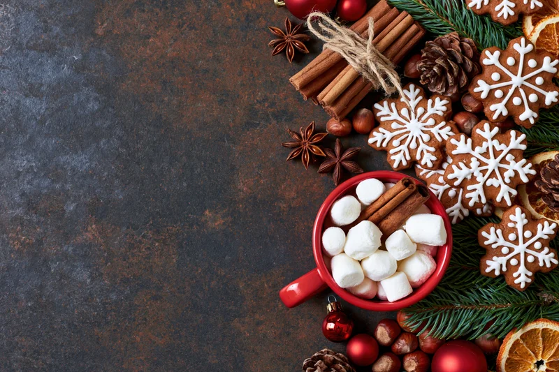 Gingerbread cookies and hot chocolate with marshmallows arranged on a dark textured surface with pine branches and holiday ornaments