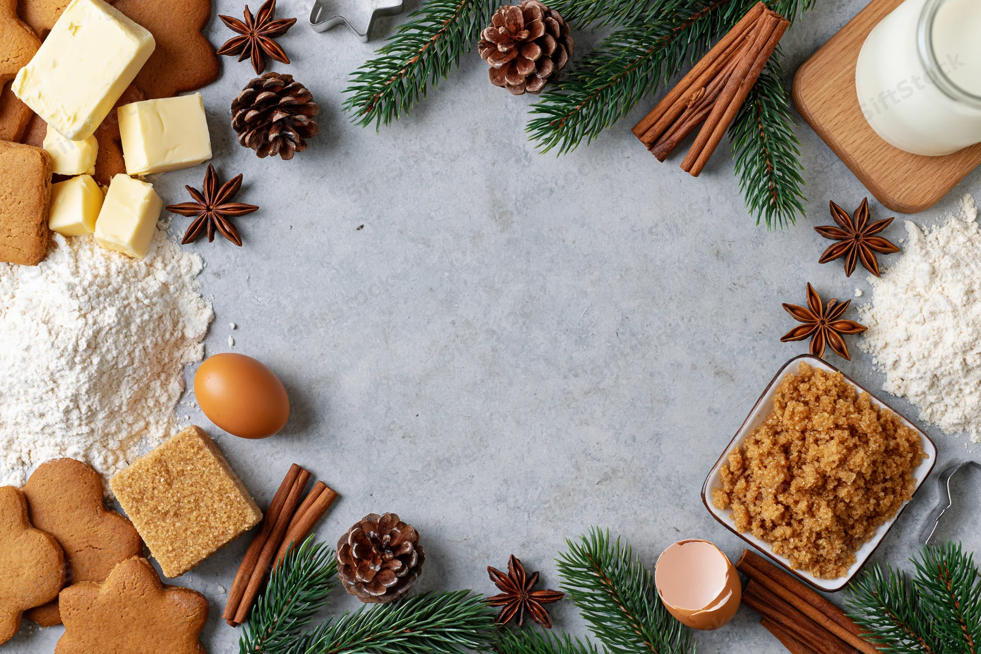 Gingerbread cookies and baking ingredients arranged around a central copyspace on a countertop with evergreen and pinecone decorations