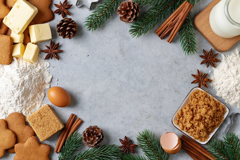 Gingerbread cookies and baking ingredients arranged around a central copyspace on a countertop with evergreen and pinecone decorations