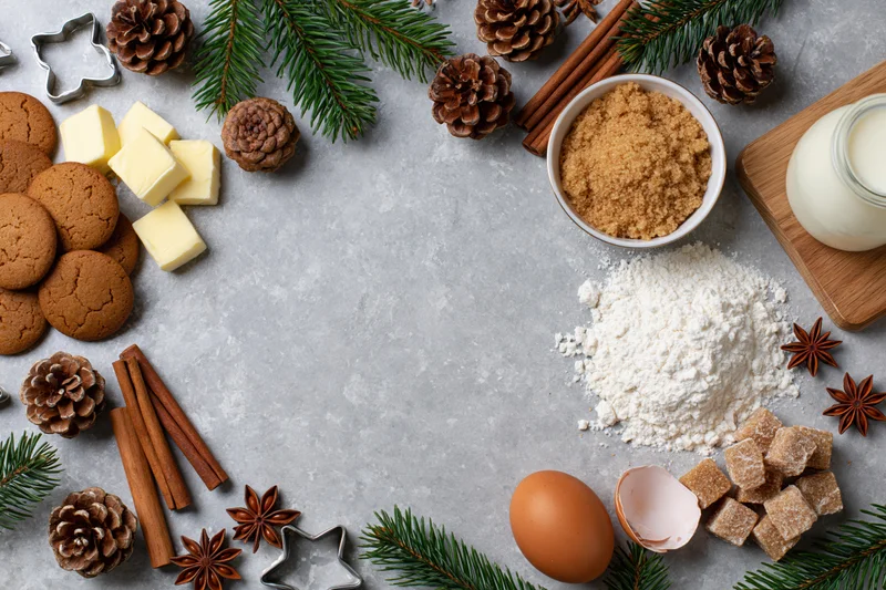 Christmas baking ingredients arranged in a festive flat lay on a gray countertop with pinecones and evergreen branches