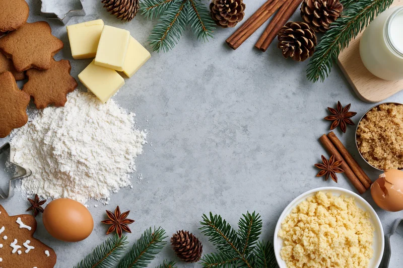 Gingerbread cookies and baking ingredients arranged around a gray countertop with evergreen branches and pinecones for christmas
