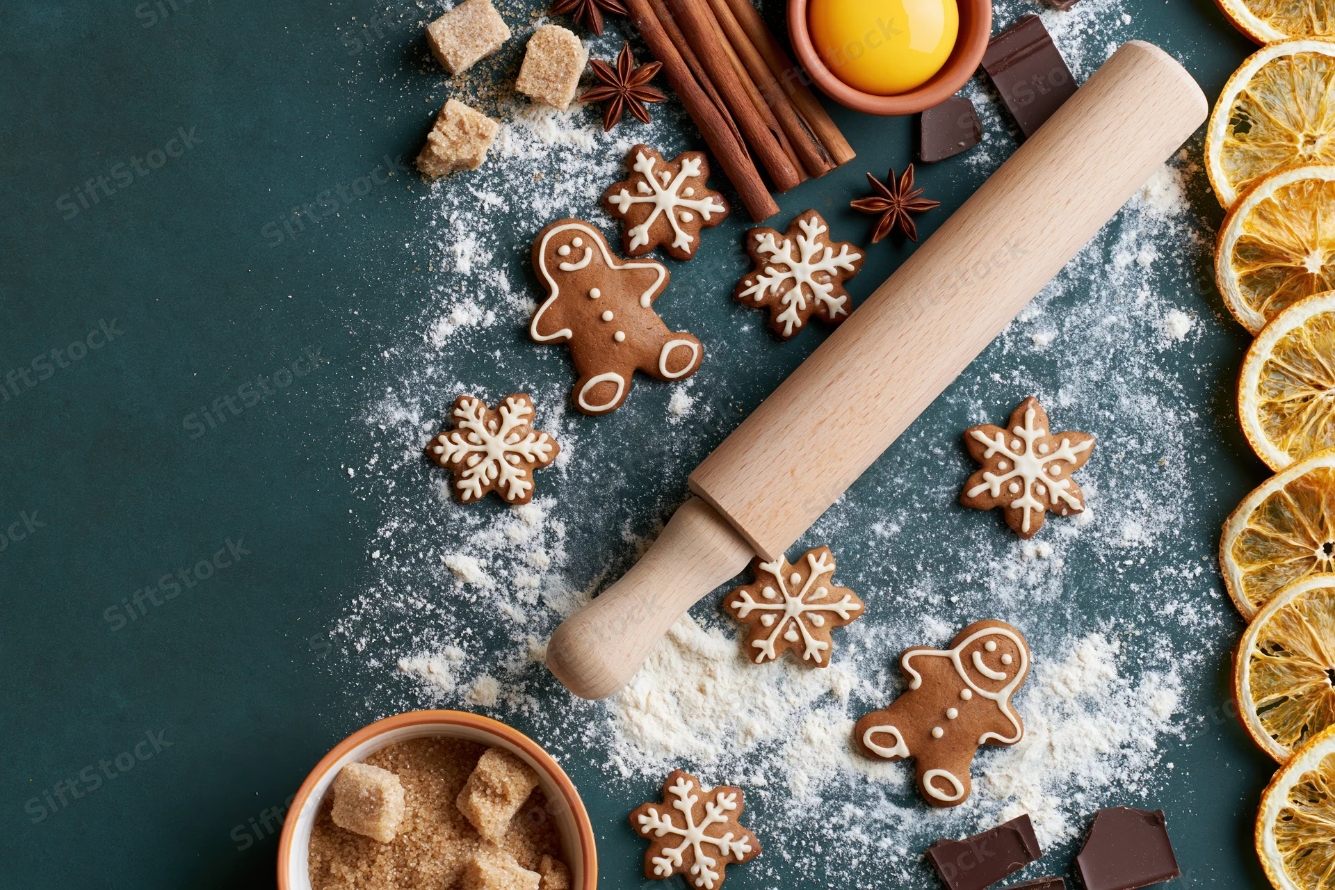 Gingerbread cookies and baking ingredients arranged on a dark surface with flour and dried oranges