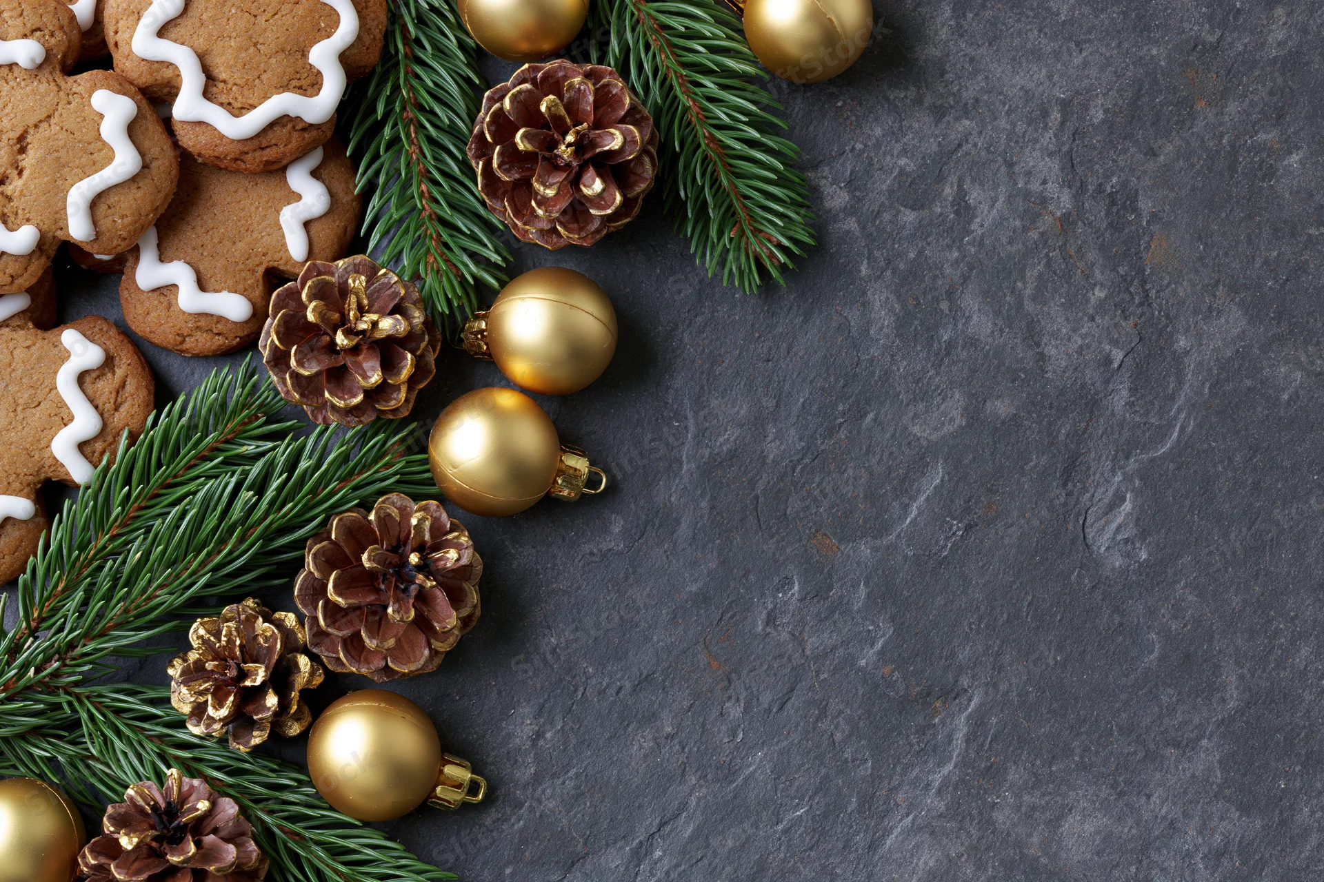 Gingerbread cookies with golden ornaments and pine branches arranged on dark slate surface