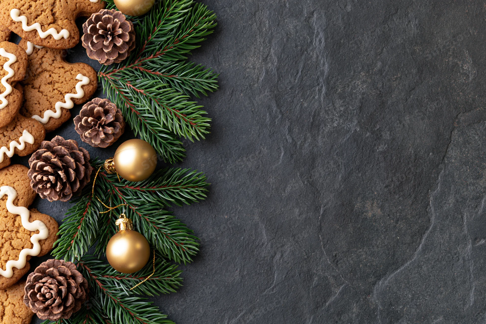 Gingerbread cookies and pine branches with ornaments arranged along the left side of a dark slate surface