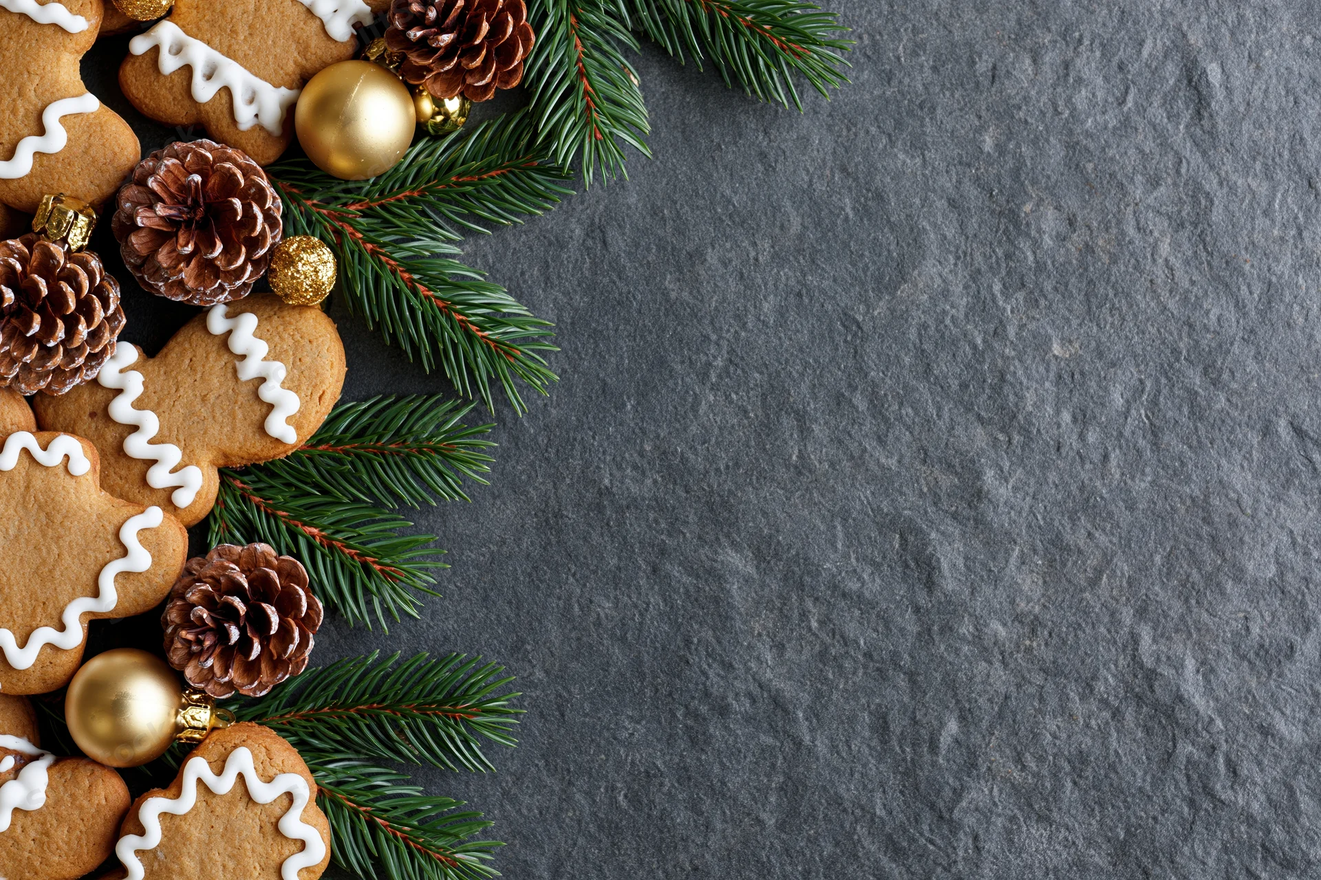 Gingerbread cookies with pine branches and ornaments arranged along the left side of a dark slate surface