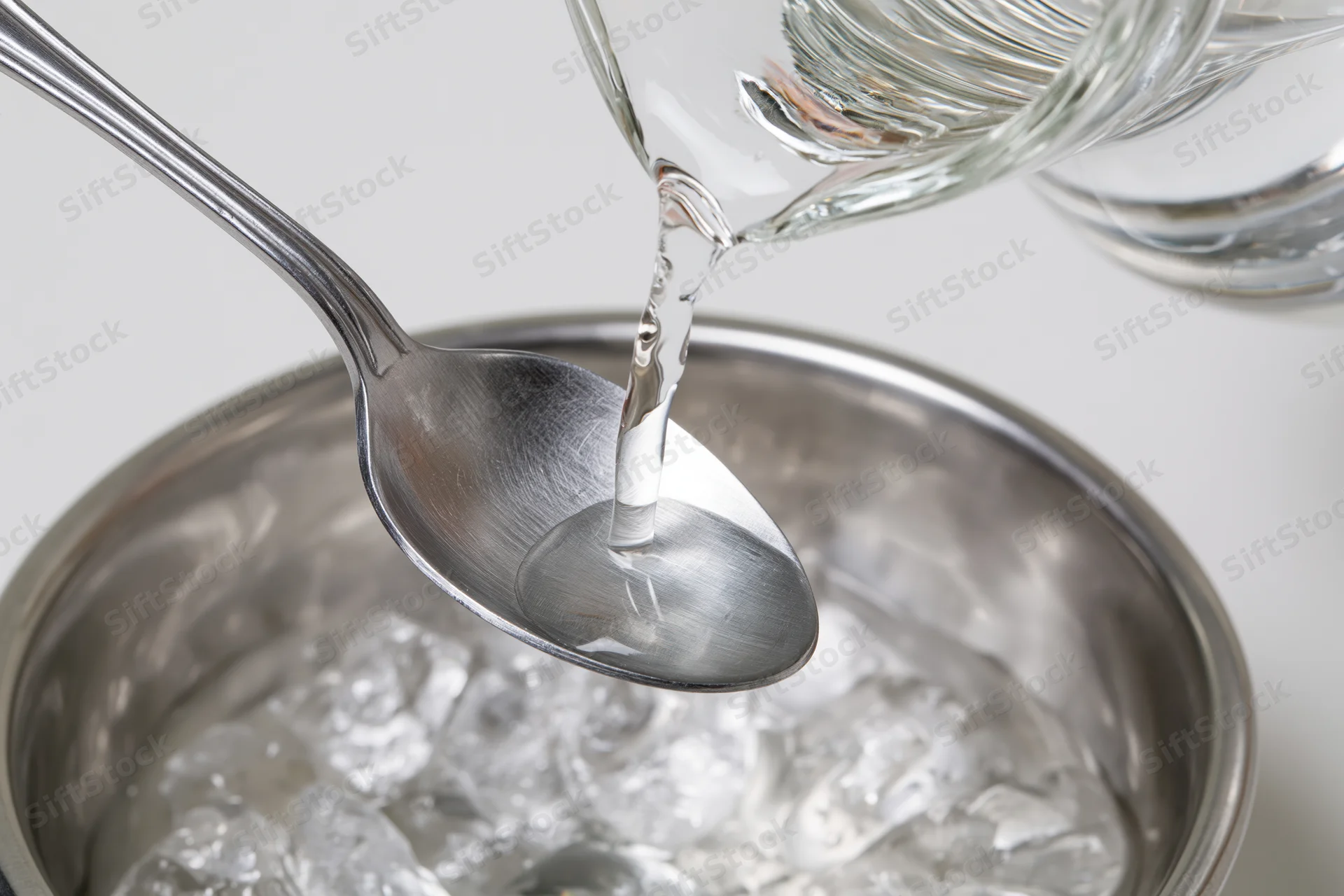 Clear liquid pouring from glass decanter into metal spoon over ice-filled bucket