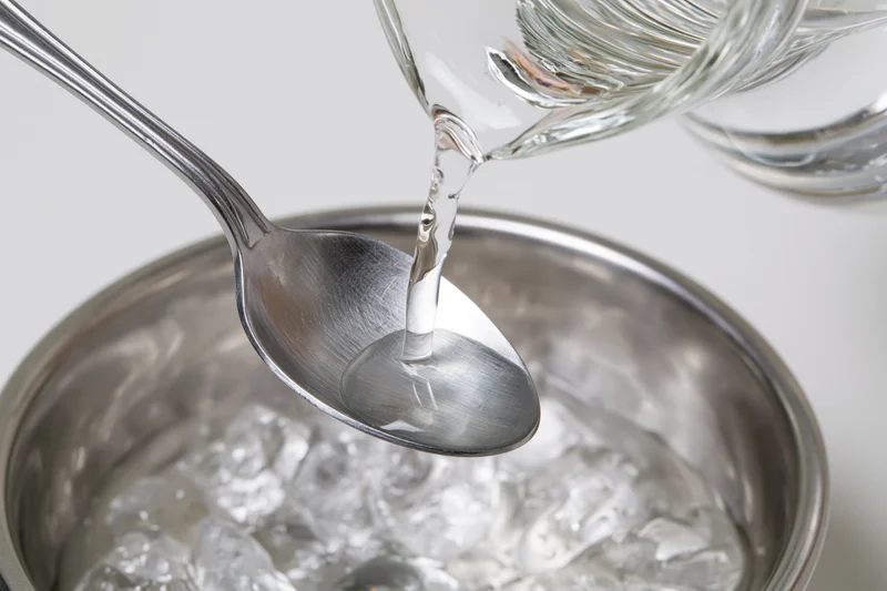 Clear liquid pouring from glass decanter into metal spoon over ice-filled bucket
