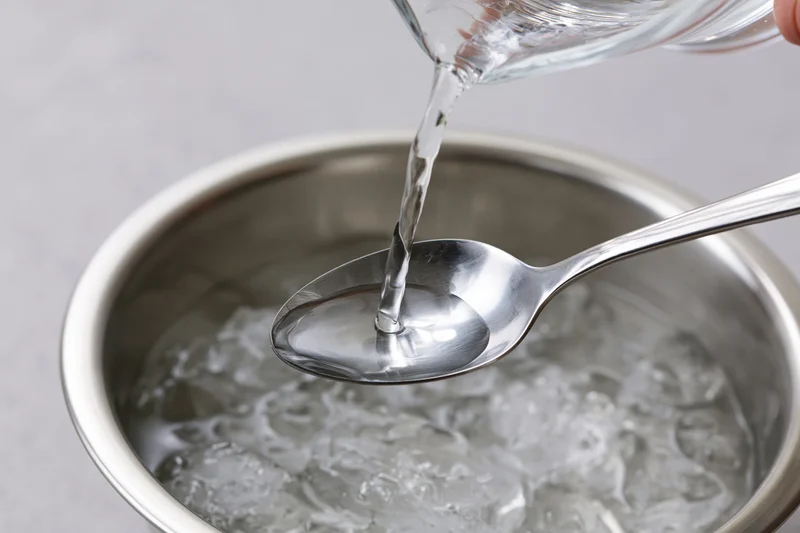 Water being poured from a glass into a spoon over a stainless steel bowl