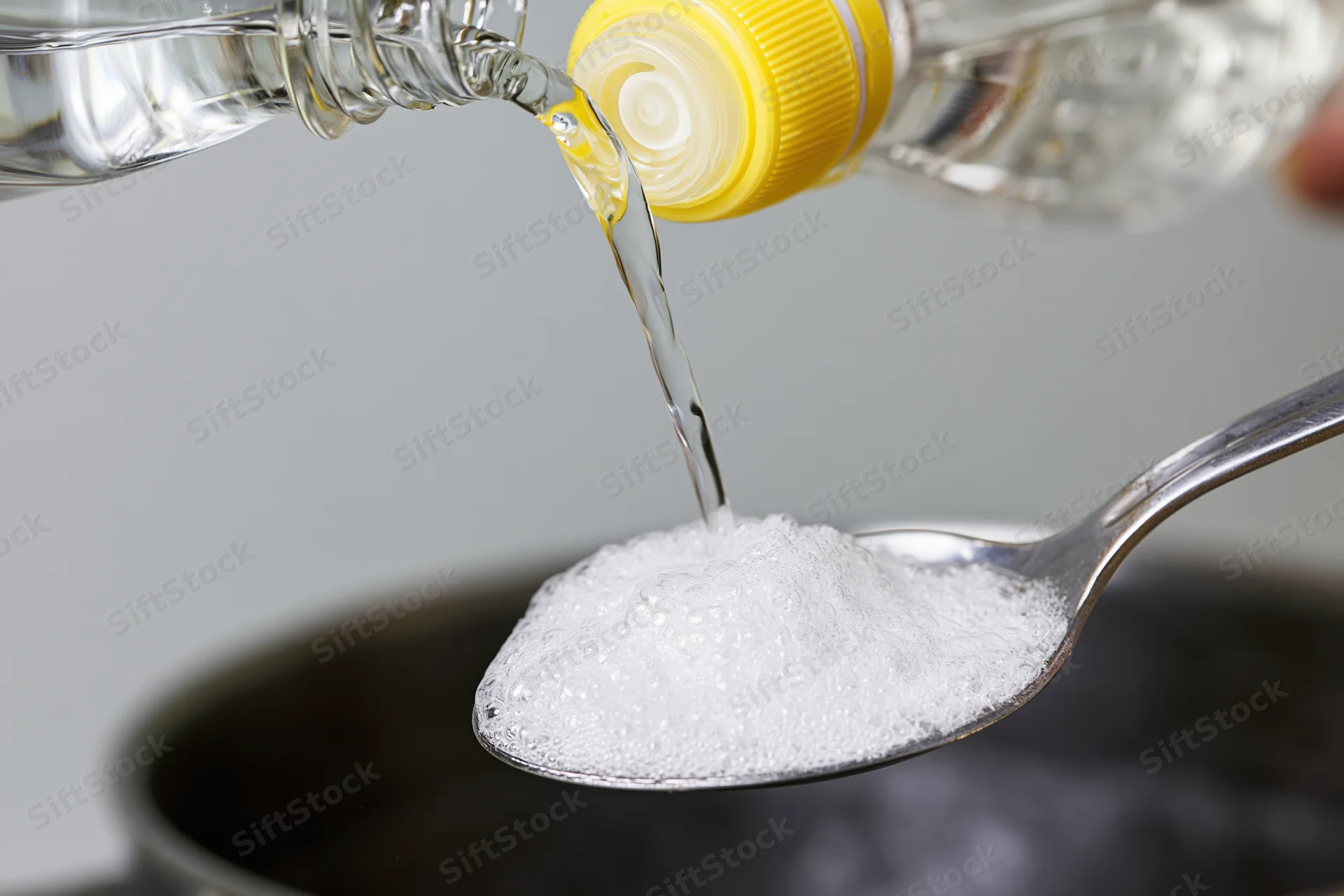 Vinegar poured from a bottle onto baking soda in a spoon, creating fizz over a dark bowl