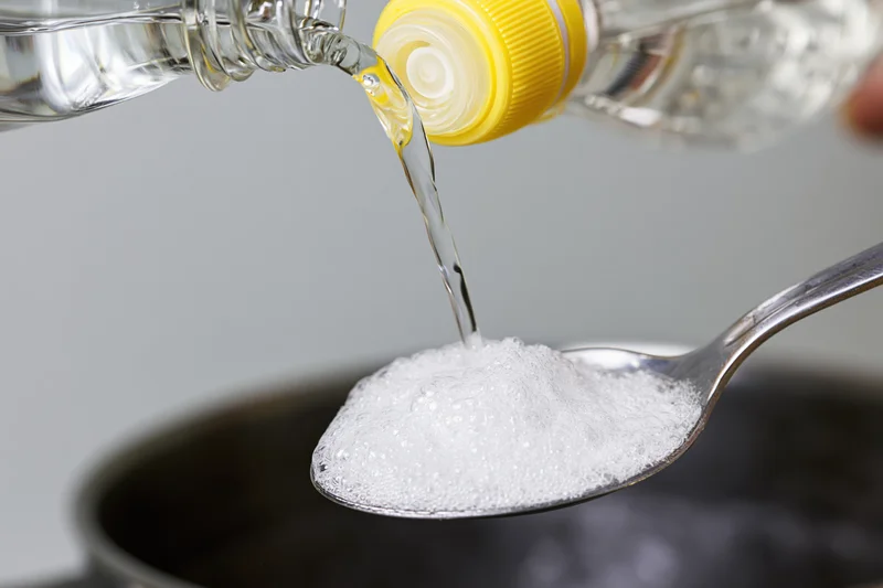 Vinegar poured from a bottle onto baking soda in a spoon, creating fizz over a dark bowl