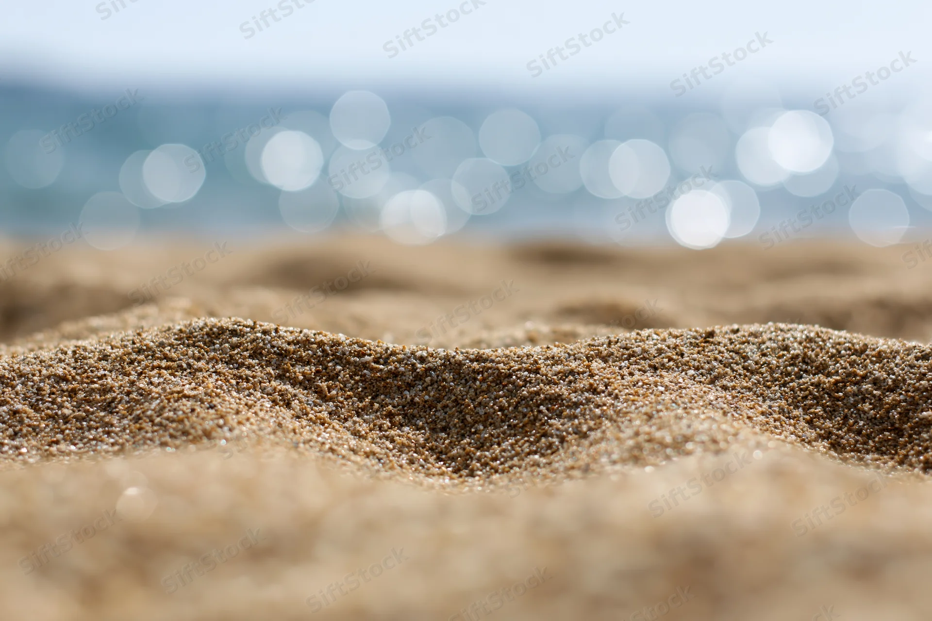 Closeup of textured sand on a beach with blurred ocean and sunlight in the background