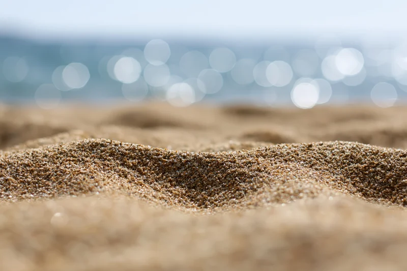 Closeup of textured sand on a beach with blurred ocean and sunlight in the background