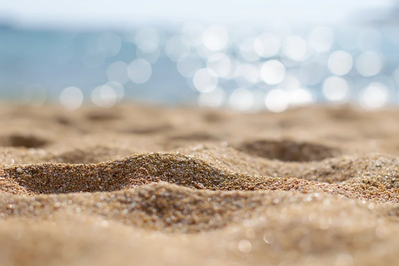 Closeup of textured sand on a beach with blurred ocean and sunlight in the background