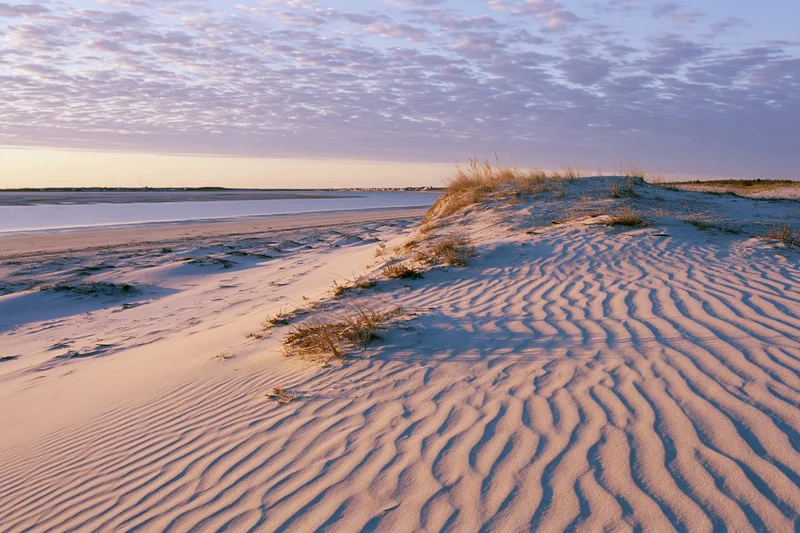 Rippled sand dune at sunset beside calm water under cloudy sky