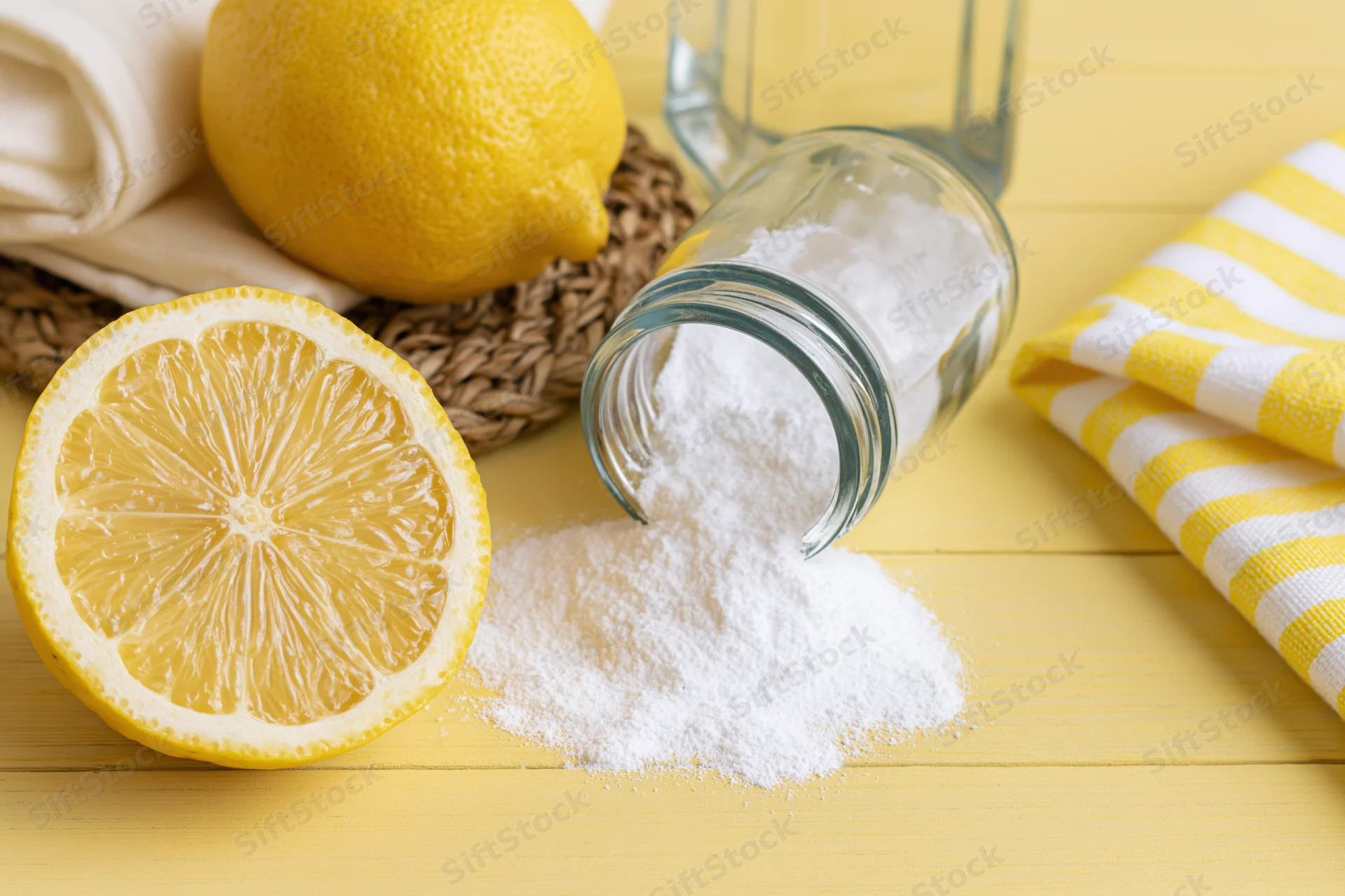 Lemon halves and spilled white powder from a glass jar on a yellow wooden surface with striped towel and woven basket