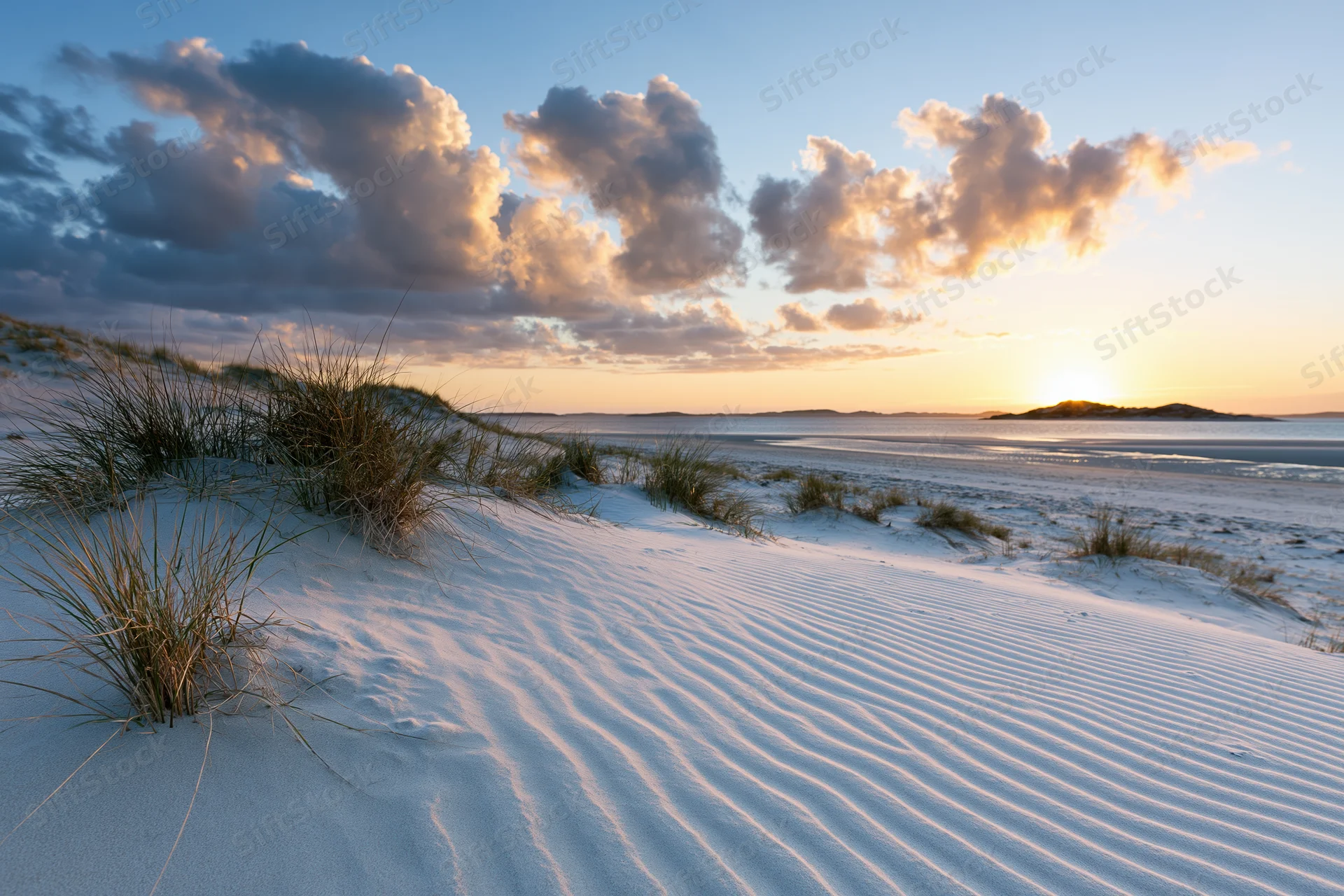 Rippled white sand dunes with beach grass under a cloudy sunset sky over the ocean