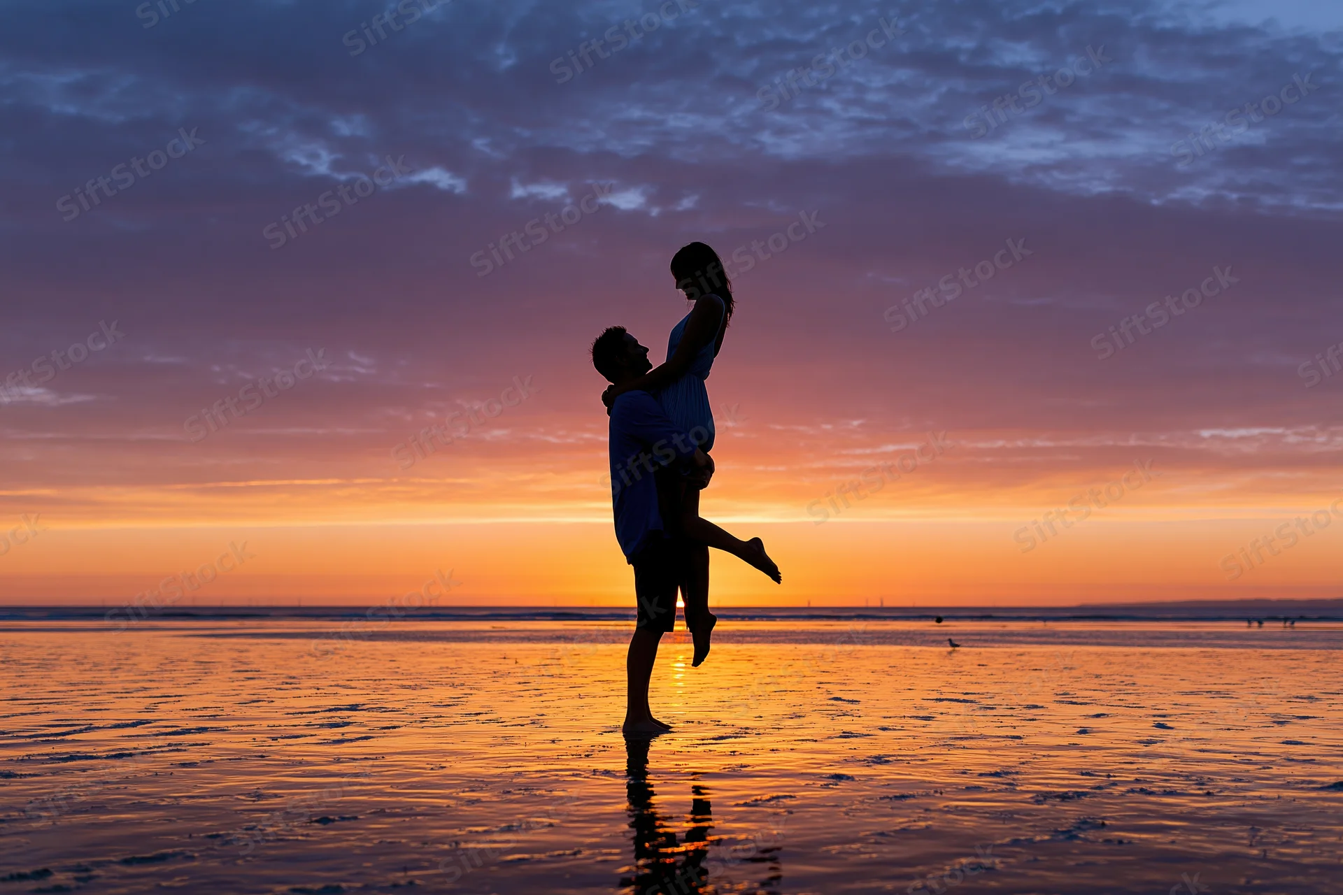Man lifting woman in silhouette against sunset sky on wet beach