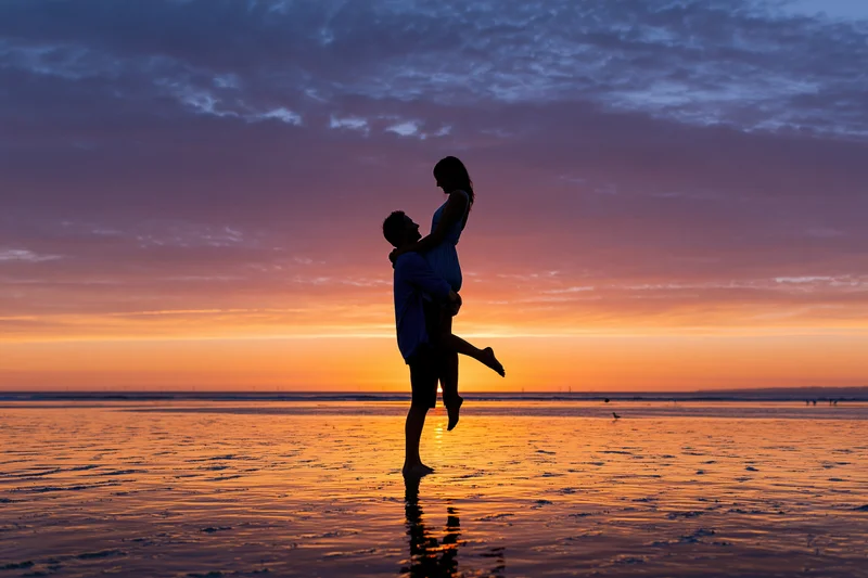 Man lifting woman in silhouette against sunset sky on wet beach