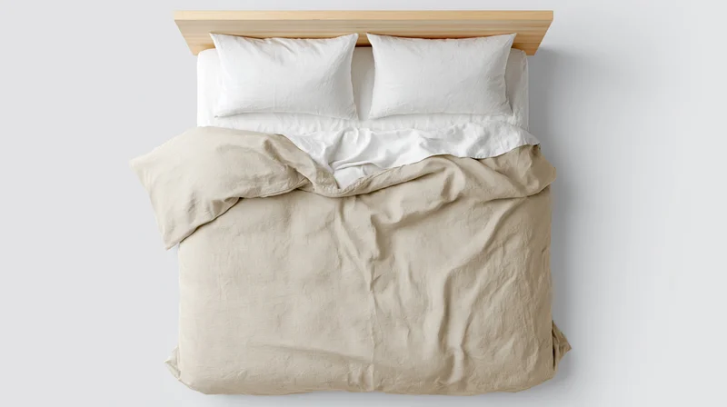 Overhead view of unmade bed with beige duvet and white pillows against plain wall