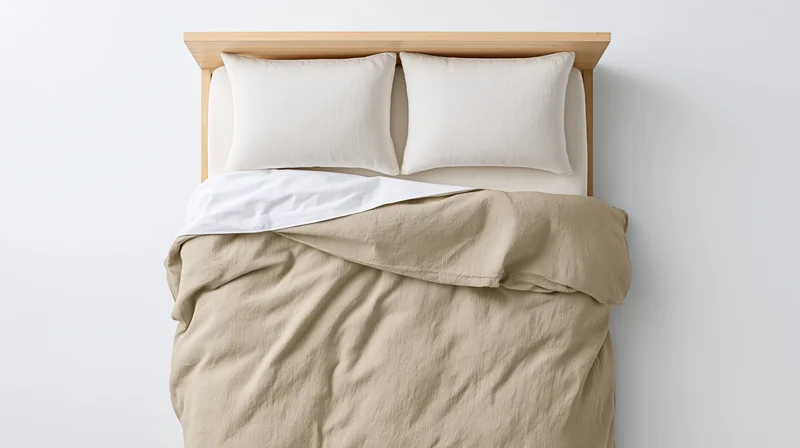 Top-down view of a neatly made bed with beige duvet and white pillows against a plain white wall