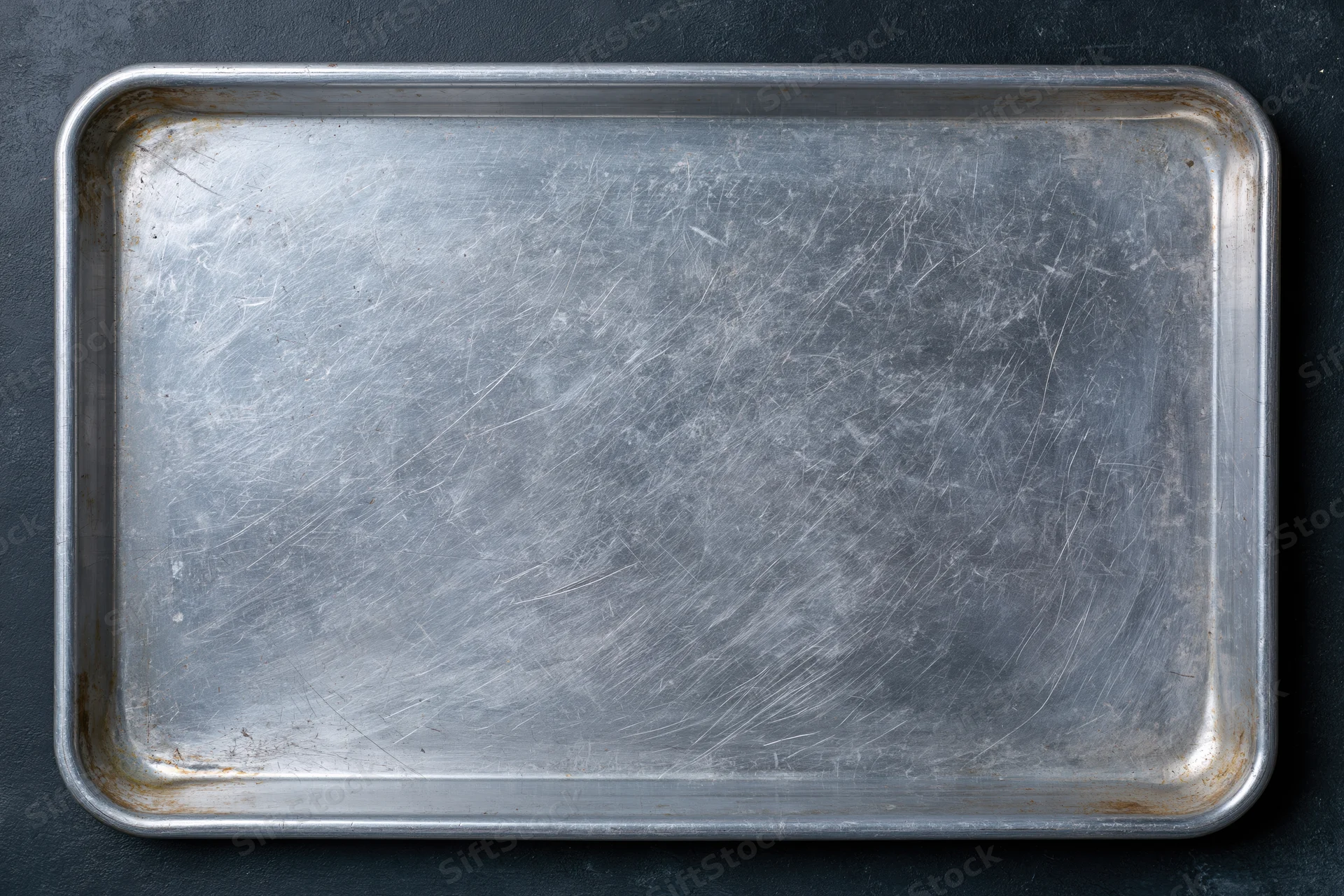 Rectangular metal baking tray with scratches and rust on dark surface, overhead view