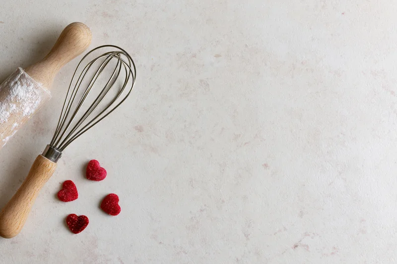 Wooden rolling pin and metal whisk with red glitter hearts on white marble countertop