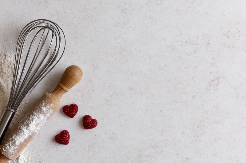 Whisk and rolling pin dusted with flour beside red glitter hearts on a light countertop