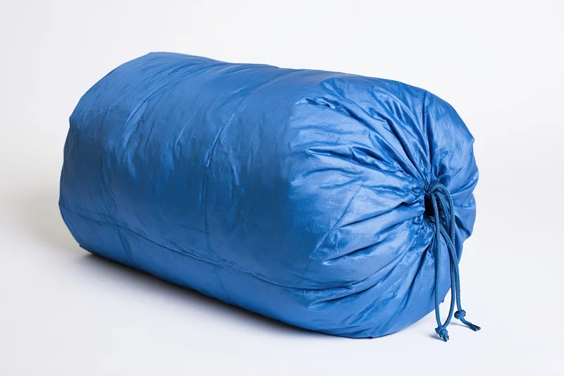 Blue nylon sleeping bag rolled and packed tightly with drawstring closure on white background