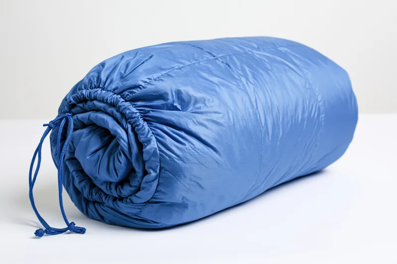 Blue nylon sleeping bag rolled and packed tightly with drawstring on white background