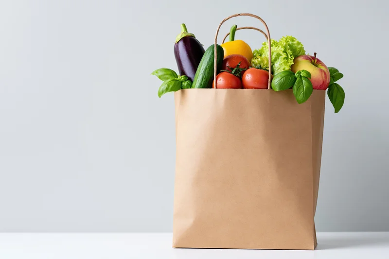 Brown paper grocery bag filled with fresh vegetables and herbs on white surface