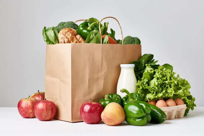 Brown paper grocery bag filled with fresh vegetables and fruits on white surface