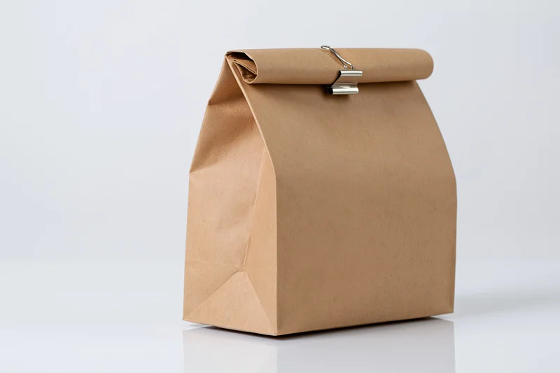 Brown kraft paper lunch bag with rolled top sealed by metal binder clip on white surface