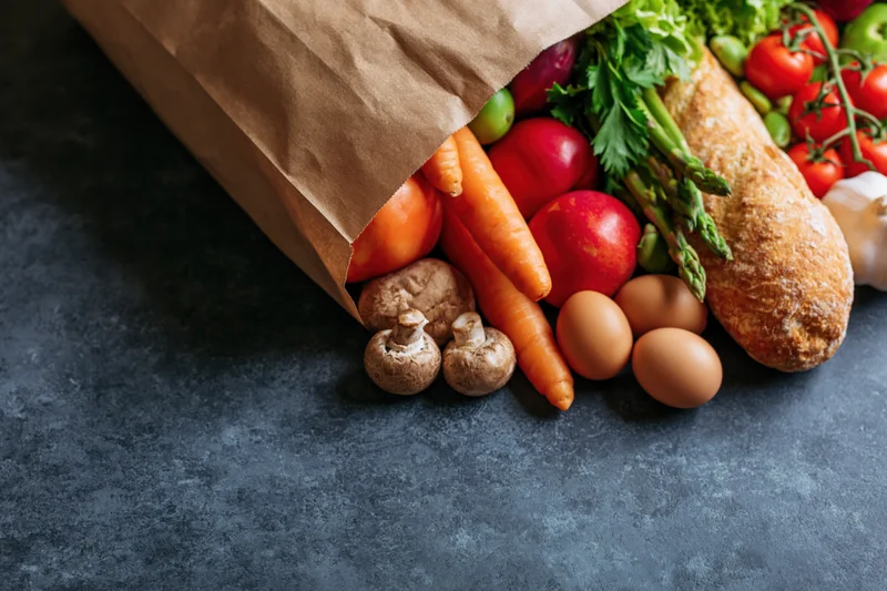 Brown paper grocery bag tipped over on dark stone surface with fresh vegetables, eggs, bread and herbs spilling out