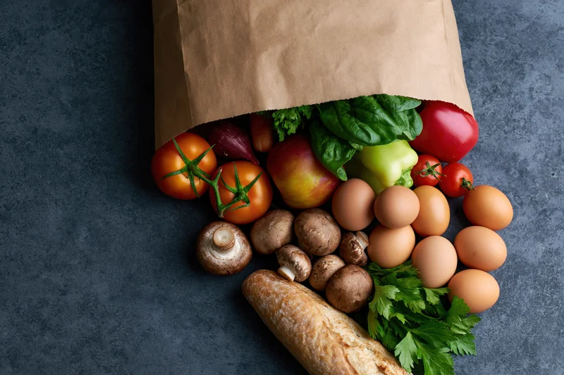 Brown paper bag spilling fresh vegetables, eggs, mushrooms, bread and herbs on dark surface