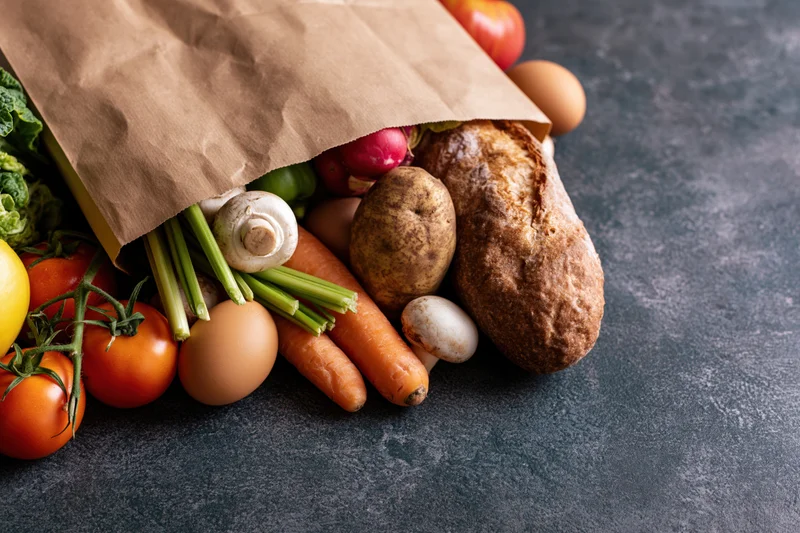 Brown paper grocery bag spilling fresh vegetables and bread on dark stone surface
