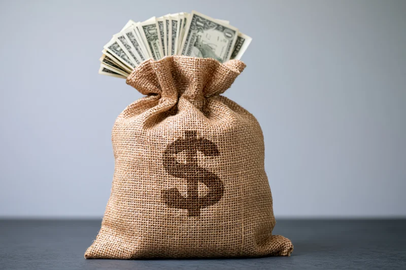 Burlap money bag with dollar sign and fanned cash bills on gray surface