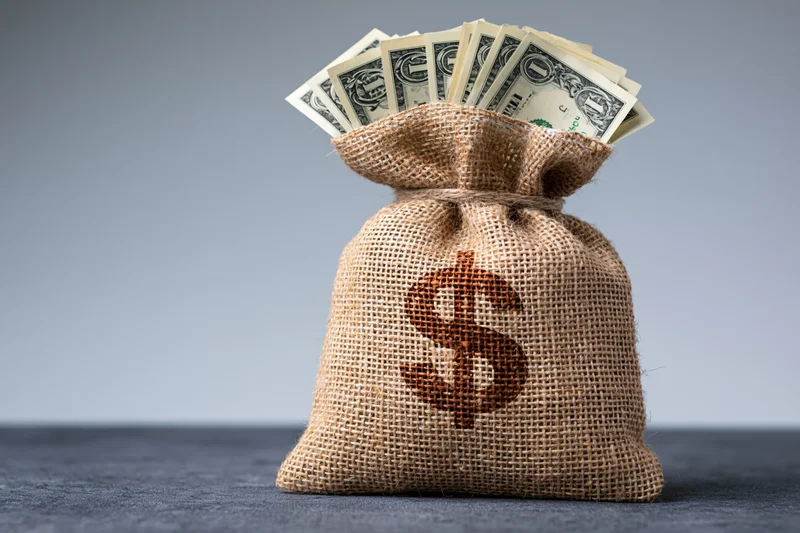 Burlap money bag with dollar sign and cash bills sticking out, on gray surface