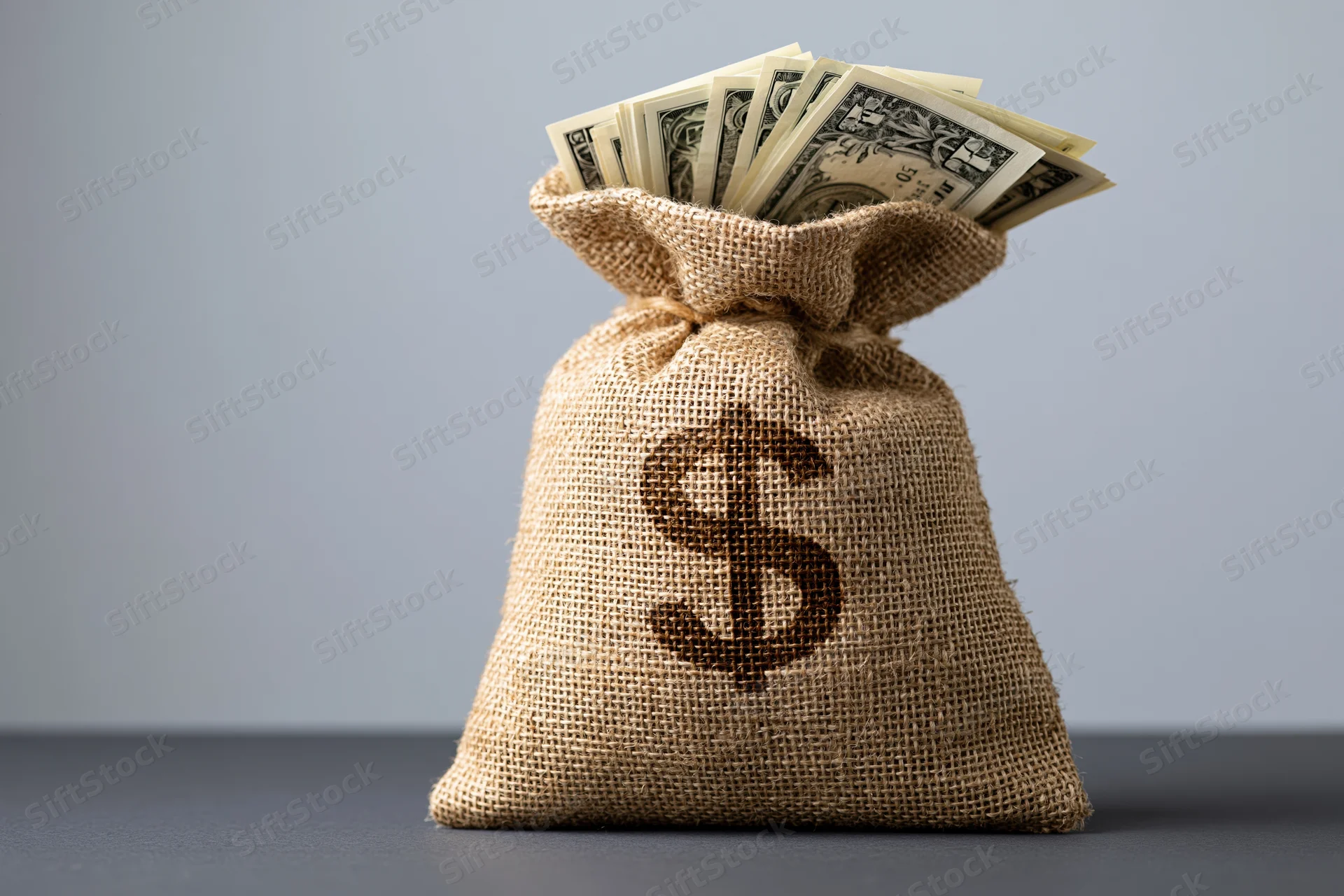 Burlap money bag with dollar sign and cash spilling out on gray surface
