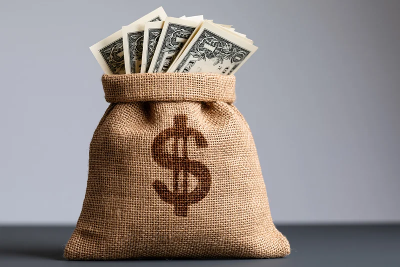 Burlap money bag with dollar sign and cash bills sitting upright on a gray surface