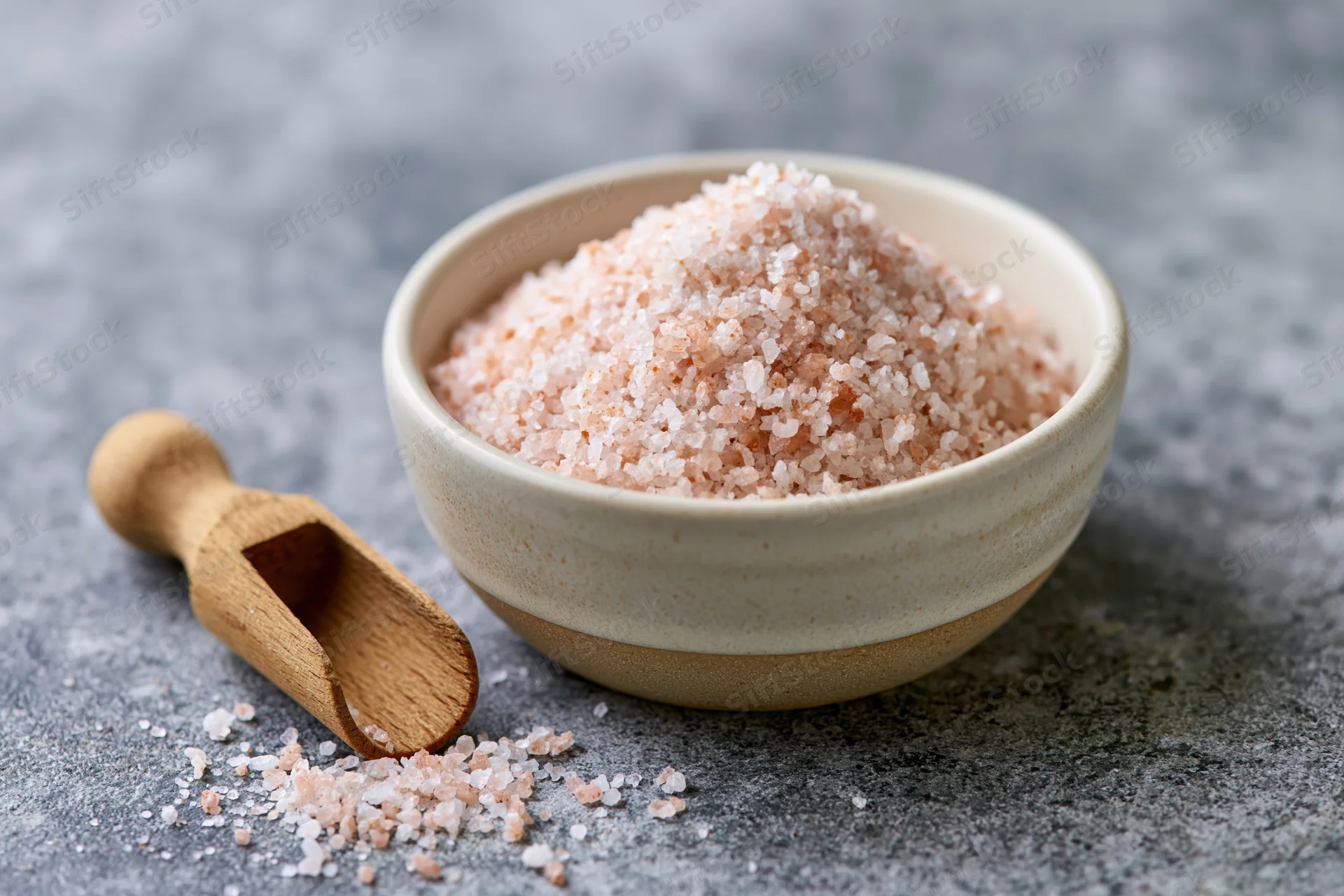 Pink himalayan salt in a ceramic bowl with wooden scoop on gray stone surface